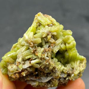 May include: A close-up of a mineral specimen featuring a cluster of light green, needle-like crystals. The crystals are clustered together, with some areas showing a brownish hue. The background is a blurred, neutral tone.