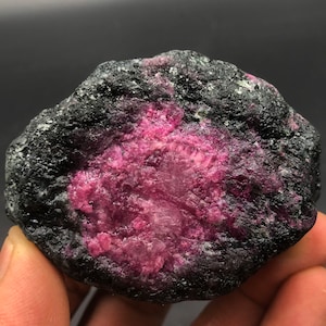 May include: A rough, dark gray rock with a large, pink ruby crystal embedded inside. The ruby has a rough, textured surface and is surrounded by the dark gray rock.