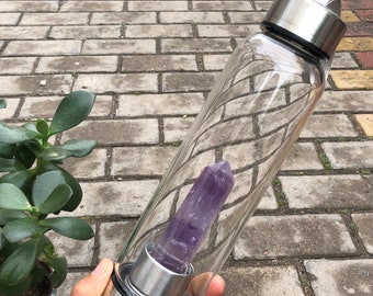 Puede incluir: Una botella de agua de vidrio transparente con una tapa de acero inoxidable y un gran cristal de amatista en su interior.