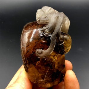 May include: A carved gemstone sculpture of a monkey atop a rounded form. The sculpture is mainly brown with translucent grey accents, displaying intricate details and a polished finish. The monkey's form is clearly defined, adding a touch of artistry.