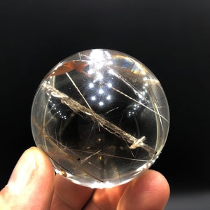 May include: A clear quartz sphere with gold and black inclusions. The sphere is approximately 5 cm in diameter and is held in a hand.