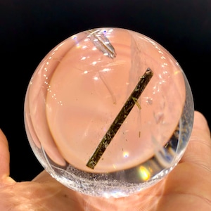 May include: A clear quartz crystal sphere with a dark green inclusion in the centre. The sphere is smooth and polished, and the inclusion is visible through the crystal.