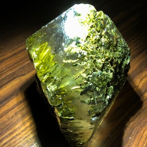 Puede incluir: Un gran cristal transparente con inclusiones verdes. El cristal tiene una forma irregular y una superficie rugosa y texturizada.