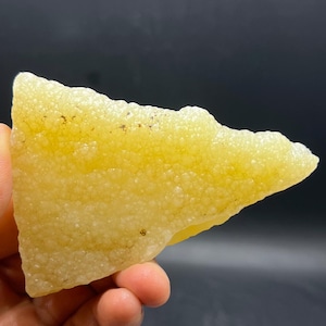 May include: A rough-edged, yellow-colored mineral specimen with a textured, bubbly surface. The mineral is held in a hand against a dark background, showcasing its irregular shape and color variations. The specimen appears to be a natural, unpolished stone.