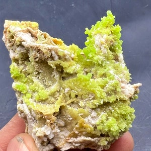 Puede incluir: Un cúmulo de cristales verde brillante sobre una roca blanca y marrón. Los cristales están agrupados en una formación densa, creando una exhibición visualmente llamativa.