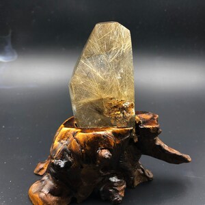 May include: A clear quartz crystal with golden rutile inclusions, presented on a carved wooden stand. The crystal is rectangular with a polished surface, and the rutile needles create a striking visual effect. The wooden stand has a natural, organic shape.