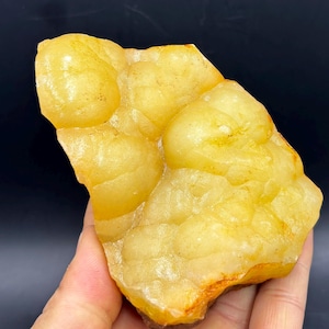May include: A raw, natural yellow calcite mineral specimen. The stone has a bumpy, rounded texture and a warm, golden-yellow color with hints of orange. The mineral is held in a hand, showcasing its size and natural form.