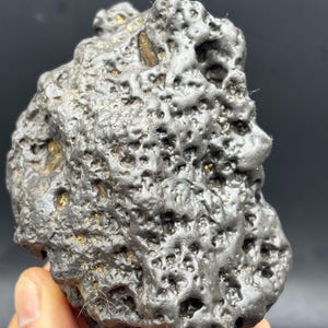 Puede incluir: Una roca oscura y porosa con una superficie rugosa y texturizada. La roca tiene una forma única con numerosos agujeros pequeños y algunas áreas con inclusiones de color dorado. La apariencia general sugiere un origen natural, posiblemente extraterrestre.