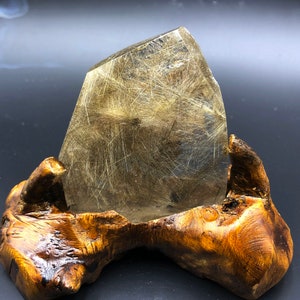 May include: A quartz crystal with golden rutile needles, presented on a carved wooden stand. The crystal is a truncated pyramid shape, highlighting the natural patterns within. The wooden stand is a warm brown colour, enhancing the crystal's display.
