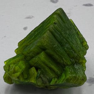 Puede incluir: Un vibrante espécimen mineral cristalino. El objeto es de un color verde lima brillante, con una estructura geométrica en capas. La superficie tiene un aspecto texturizado, con crestas y facetas visibles. La forma general es aproximadamente piramidal, con múltiples capas apiladas.