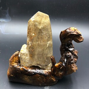 May include: A clear quartz crystal with golden inclusions, mounted on a dark brown wooden base. The crystal is rectangular with a pointed top. The wooden base has a natural, organic form. The crystal is a decorative item.
