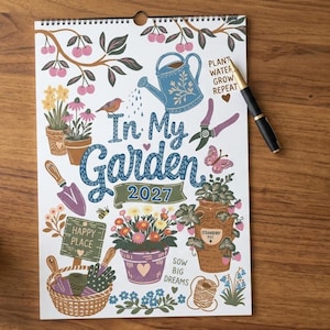 Könnte beinhalten: Ein Wandkalender für 2027 mit dem Titel In My Garden, der bunte Illustrationen einer blauen Gießkanne, Topfblumen, eines Erdbeertopfs, eines Korbs mit Werkzeugen, eines Vogels, eines Schmetterlings sowie den Text Plant Water Grow Repeat und Sow Big Dreams zeigt.