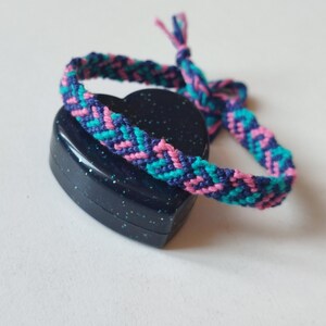 May include: A colourful friendship bracelet in teal, pink, and purple, resting on a dark blue heart-shaped box. The bracelet has a chevron pattern and is tied at the end.