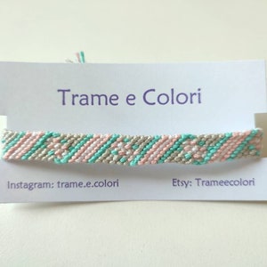 Op de afbeelding: Handgemaakte vriendschapsarmband in pastelkleuren roze, mintgroen en beige. De geweven armband wordt gepresenteerd op een witte kaart met de tekst "Trame e Colori", Instagram- en Etsy-accountinformatie.