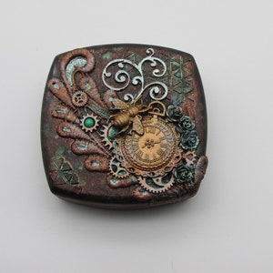 Könnte beinhalten: Eine kleine, quadratische, dunkelbraune Schachtel mit einem dekorativen Design mit einer goldenen Biene, einem Zifferblatt, Zahnrädern und grünen Akzenten. Die Schachtel hat eine Steampunk-Ästhetik.