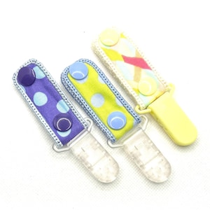 May include: Three fabric pacifier clips with plastic fasteners. The first is purple with light blue polka dots and dark purple snaps. The second is lime green with light blue polka dots and light blue snaps. The third is yellow with a multi-colored pattern.