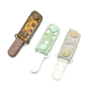 May include: Three baby pacifier clips in a top-down view. One clip has a brown giraffe print, another has a green floral print, and the last is a solid gray. Each clip has a plastic clasp and two buttons.