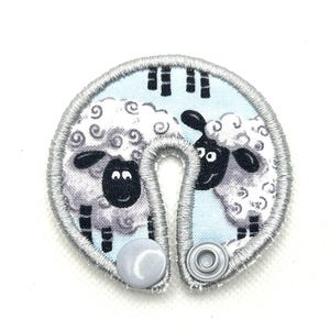May include: A circular fabric cord wrap featuring a light blue background with a repeating pattern of white and black sheep. The edges are finished with silver-grey embroidery stitching, and the piece includes two silver-colored plastic snap fasteners.