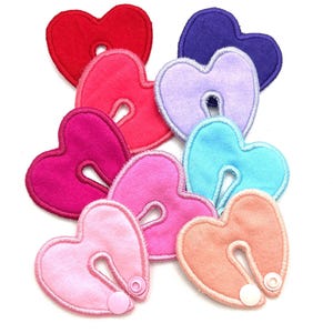May include: A collection of colorful heart-shaped appliqués in various shades of red, pink, purple, and blue. Each heart has a unique design with a central opening and a snap closure. The appliqués are made of soft fabric and are ideal for crafting projects.