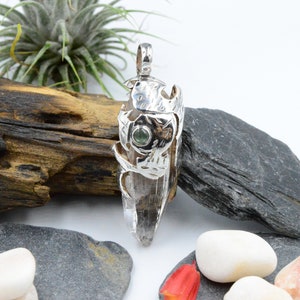 Peut inclure: Un pendentif en argent avec un cristal de quartz clair enchâssé dans un design argenté détaillé. Une petite pierre précieuse verte est sertie dans le cadre en argent.