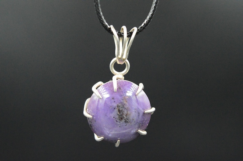 K&ouml;nnte beinhalten: Ein runder, violetter Anh&auml;nger mit einer silberfarbenen Metalleinfassung und einer schwarzen Kordel. Der Anh&auml;nger hat eine polierte, glatte Oberfl&auml;che mit dunklen Einschl&uuml;ssen. Die Metalleinfassung hat ein dekoratives, offenes Design.