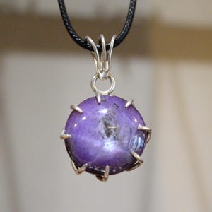 K&ouml;nnte beinhalten: Ein runder, lila Edelsteinanh&auml;nger mit einer silberfarbenen Metalleinfassung und einer schwarzen Kordel. Der Edelstein hat ein sternf&ouml;rmiges Muster in der Mitte. Der Anh&auml;nger h&auml;ngt vor einem verschwommenen Hintergrund.