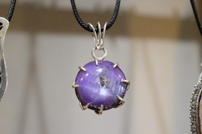 K&ouml;nnte beinhalten: Ein runder, lila Edelsteinanh&auml;nger mit einer silberfarbenen Metalleinfassung und einer schwarzen Kordel. Der Edelstein hat ein sternf&ouml;rmiges Muster in der Mitte. Der Anh&auml;nger h&auml;ngt vor einem verschwommenen Hintergrund.