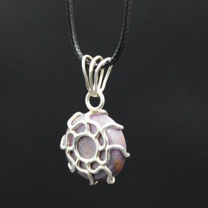 K&ouml;nnte beinhalten: Eine Anh&auml;ngerkette mit einem runden, lavendelfarbenen Stein, der in eine silberfarbene, netzartige Fassung gefasst ist. Der Anh&auml;nger h&auml;ngt an einem schwarzen Band vor dunklem Hintergrund. Ein einzigartiges Schmuckaccessoire.