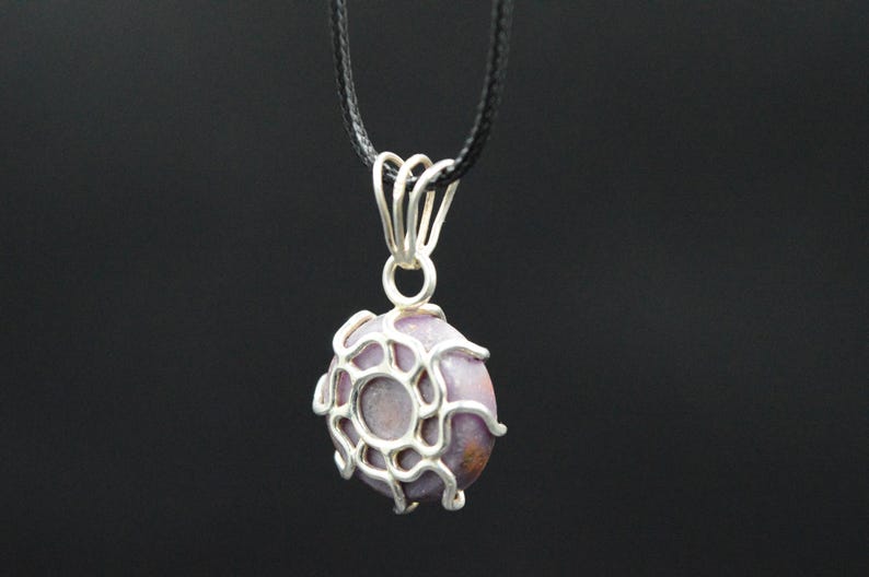 K&ouml;nnte beinhalten: Eine Anh&auml;ngerkette mit einem runden, lavendelfarbenen Stein, der in eine silberfarbene, netzartige Fassung gefasst ist. Der Anh&auml;nger h&auml;ngt an einem schwarzen Band vor dunklem Hintergrund. Ein einzigartiges Schmuckaccessoire.