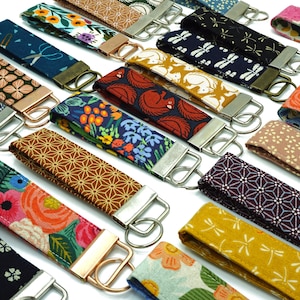 Könnte beinhalten: Eine Sammlung von Schlüsselanhängern aus Stoff mit verschiedenen floralen, geometrischen und Tiermustern. Die Schlüsselanhänger verfügen über Metallbeschläge und eine Schlaufe zum Befestigen von Schlüsseln. Farben sind Blau, Orange, Gelb und Rosa.