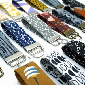 May include: A collection of colourful fabric keychains with different patterns and designs. Each keychain has a metal ring and a silver or bronze clasp.