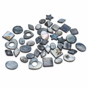 Puede incluir: Una colección de cuentas de concha iridiscentes, gris-azuladas, en varias formas, incluyendo cuadrados, diamantes, corazones, estrellas y óvalos. Algunas cuentas tienen un brillo nacarado y patrones naturales. Las cuentas están dispersas sobre una superficie blanca.