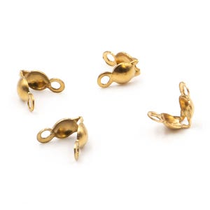 May include: Four gold-coloured metal crimp covers, each featuring a rounded, hollow shell design with two small loops extending from opposite sides. These jewellery findings are arranged in a scattered pattern against a plain white background.