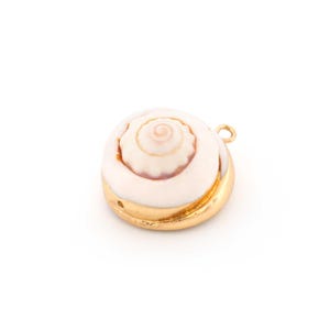 Könnte beinhalten: Ein runder, weißer Muschelanhänger mit Spiralmuster, eingefasst in einen goldfarbenen Metallrahmen. Der Anhänger hat eine kleine Schlaufe zum Befestigen an einer Kette oder einer anderen Schmuckkomponente. Die Muschel hat ein natürliches, organisches Aussehen.