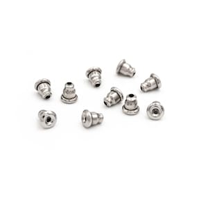 Pode incluir: Uma coleção de fechos de brincos prateados espalhados sobre uma superfície branca. Os fechos têm uma forma arredondada, semelhante a um sino, com um pequeno orifício no centro. O metal tem uma qualidade brilhante e refletora.
