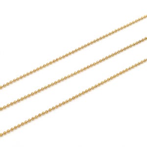 Könnte beinhalten: Eine goldfarbene Kugelkette, die aus einer Reihe kleiner, gleichmäßiger, kugelförmiger Perlen besteht, die durch dünne Metallglieder verbunden sind. Die Kette ist in drei parallelen, diagonalen Linien vor einem weißen Hintergrund dargestellt.