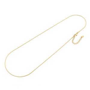 Könnte beinhalten: Eine goldfarbene Schlangenkette mit glattem, flexiblem Design. Die Halskette verfügt über einen Karabinerverschluss und eine verstellbare Verlängerungskette mit einem kleinen rechteckigen Metallanhänger am Ende für einen sicheren Sitz.