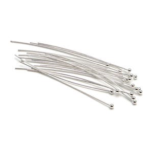 Puede incluir: Una colección de alfileres de cabeza de metal de color plateado, cada uno con un vástago largo, fino y recto y una pequeña bola esférica en un extremo. Estos componentes para joyería están dispuestos en un grupo suelto y superpuesto.