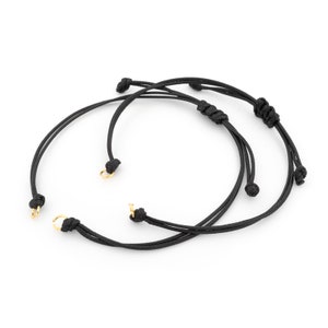 Könnte beinhalten: Zwei schwarze Kordelarmbänder mit goldenen Metallverschlüssen. Die Armbänder sind an den Enden verknotet.
