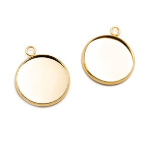May include: Two circular gold-toned metal pendants with a smooth, flat surface and a raised rim. Each pendant features a small, integrated circular loop at the top for attachment, designed for jewellery making or decorative craft projects.