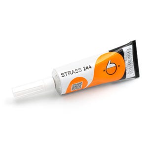 May include: A white squeeze tube of Strass 244 adhesive featuring orange circular graphics, a black end seal, a QR code, and a white plastic cap. The label displays the text Strass 244 along with a barcode and product identification numbers.