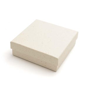 Könnte beinhalten: Eine quadratische Geschenkbox mit Deckel aus cremefarbenem, gesprenkeltem Papier. Die Box besteht aus zwei Teilen mit einem abnehmbaren Deckel, der auf das Unterteil passt und eine texturierte, gefleckte Oberfläche auf der Außenseite aufweist.
