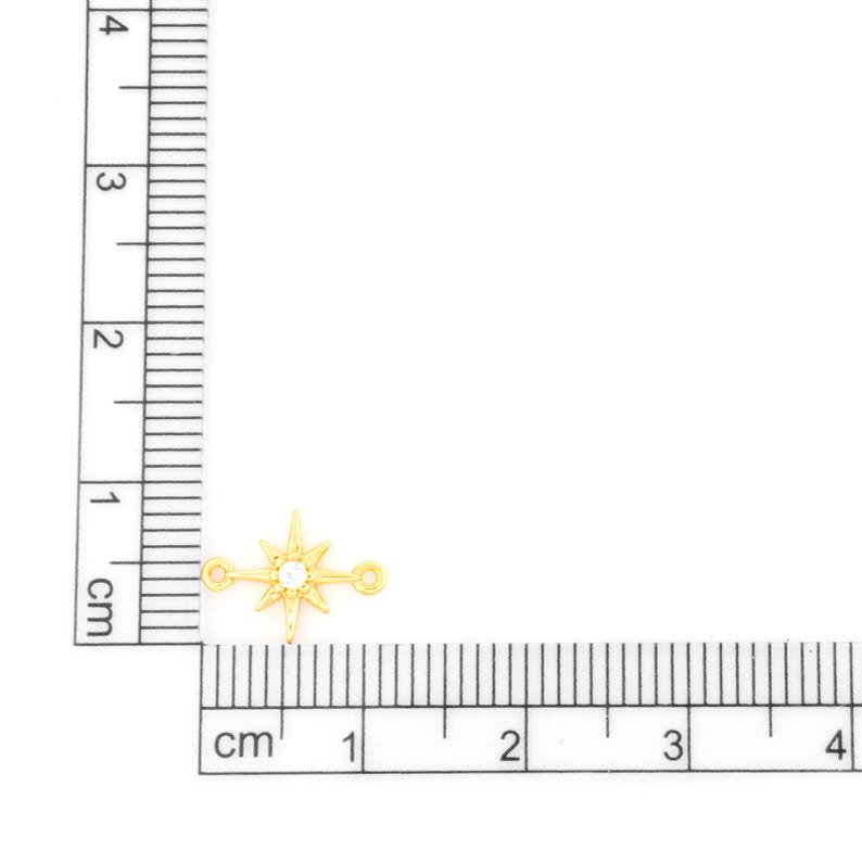 May include: A small, gold-colored charm in the shape of a compass rose, with a clear, round gem at its center. The charm has two small loops on either side. The image includes a ruler with centimeter markings.