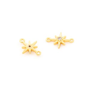 May include: Two gold-colored starburst charms with small loops on each end for jewelry making. One charm has a clear crystal in the center. The charms are on a white background.