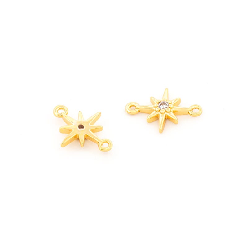 May include: Two gold-colored starburst charms with small loops on each end for jewelry making. One charm has a clear crystal in the center. The charms are on a white background.