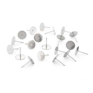 Könnte beinhalten: Eine Ansammlung silberfarbener Ohrstecker mit flachen, runden Pads. Die Stifte sind auf einer weißen Oberfläche verstreut und zeigen ihr schlichtes Design. Diese Komponenten sind ideal für die DIY-Schmuckherstellung.