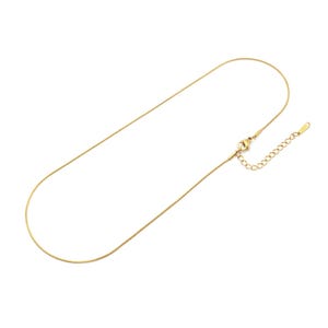 Könnte beinhalten: Eine goldfarbene Schlangenkette mit einem glatten, flexiblen Design. Die Kette verfügt über einen Karabinerverschluss und eine verstellbare Verlängerungskette mit kleinen runden Gliedern, die eine individuelle Anpassung um den Hals ermöglicht.