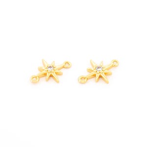 May include: Two gold-colored starburst charms with a clear crystal in the center. Each charm has two small loops for attaching to jewelry. The charms are on a white background.