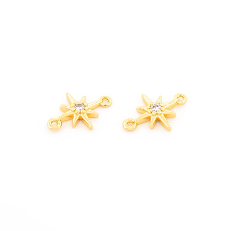 May include: Two gold-colored starburst charms with a clear crystal in the center. Each charm has two small loops for attaching to jewelry. The charms are on a white background.