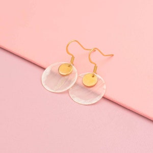 Könnte beinhalten: Ein Paar goldene Ohrringe mit einer weißen, muschelförmigen Scheibe und einer kleineren goldenen Scheibe obenauf. Die Ohrringe liegen auf einem rosafarbenen Hintergrund.